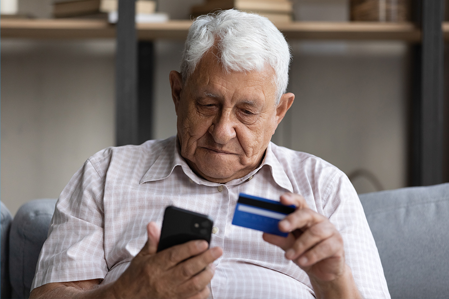 An elderly man sits on a couch, holding a credit card in one hand and looking closely at his smartphone
