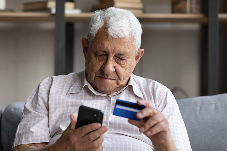 An elderly man sits on a couch, holding a credit card in one hand and looking closely at his smartphone