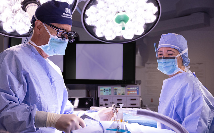 Two surgeons in blue scrubs, masks, and surgical caps perform a cardiac catheterization procedure together in an operating room